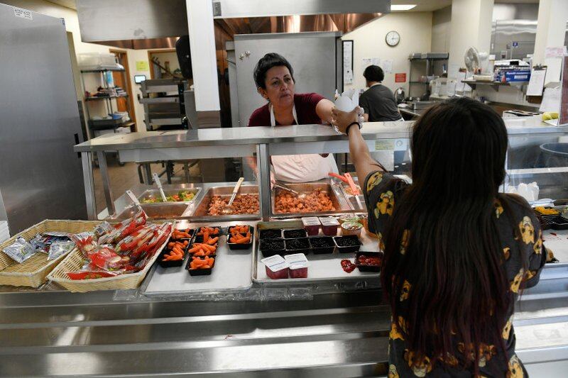 A woman food worker wearing a white apron hands hands a high schooler with long hair a take-out carton from a lunch line, with a kitchen in the background. The worker serves from pans of lunch items, while in the foreground trays feature small containers of carrots, cookies and fruit and other items to grab.