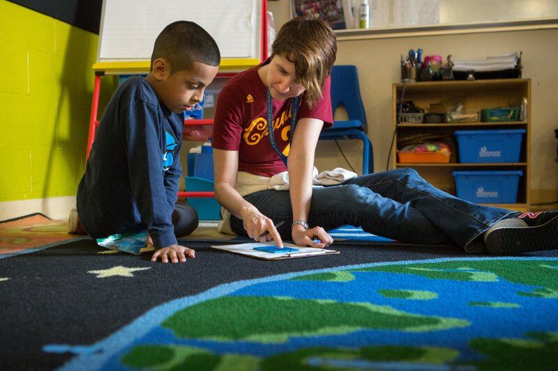 A teacher and a student sit on a rug shaped like the Earth. The teacher is pointing to a piece of paper and the student is looking at it.