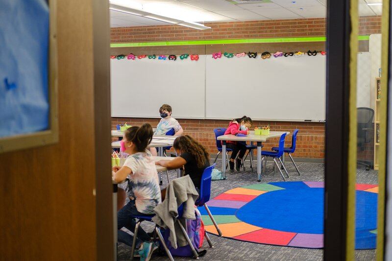 Students sit at tables in a classroom, with a colorful circle-shaped rug dominating the photo.